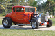 An orange hot rod is parked on the grass.